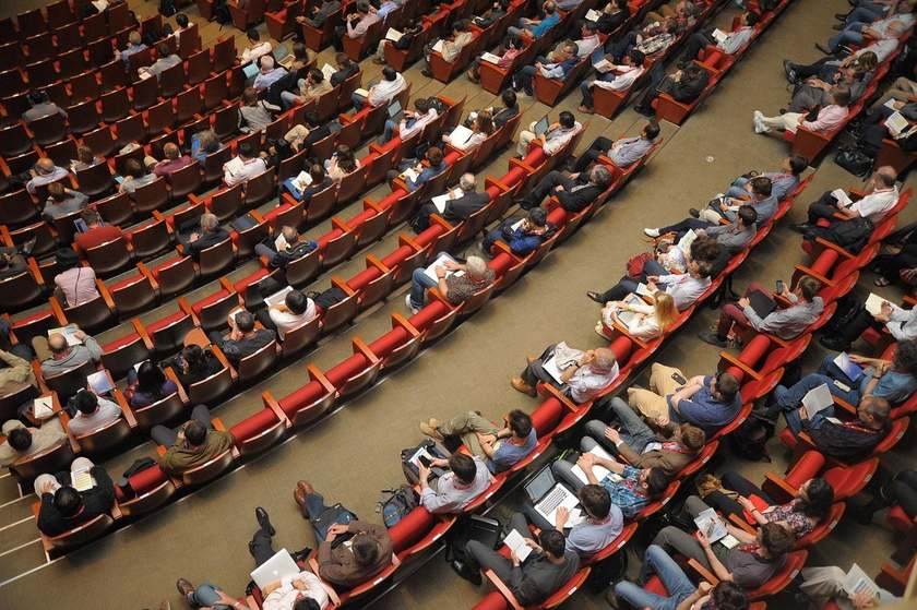 Salle de conférence avec personnes assises sur des sièges rouges, certaines utilisent des ordinateurs portables Salle de conférence avec personnes assises sur des sièges rouges, certaines utilisent des ordinateurs portables