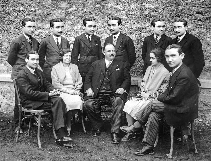 Groupe de personnes en tenue formelle, assis et debout sur des chaises, devant un mur de pierre